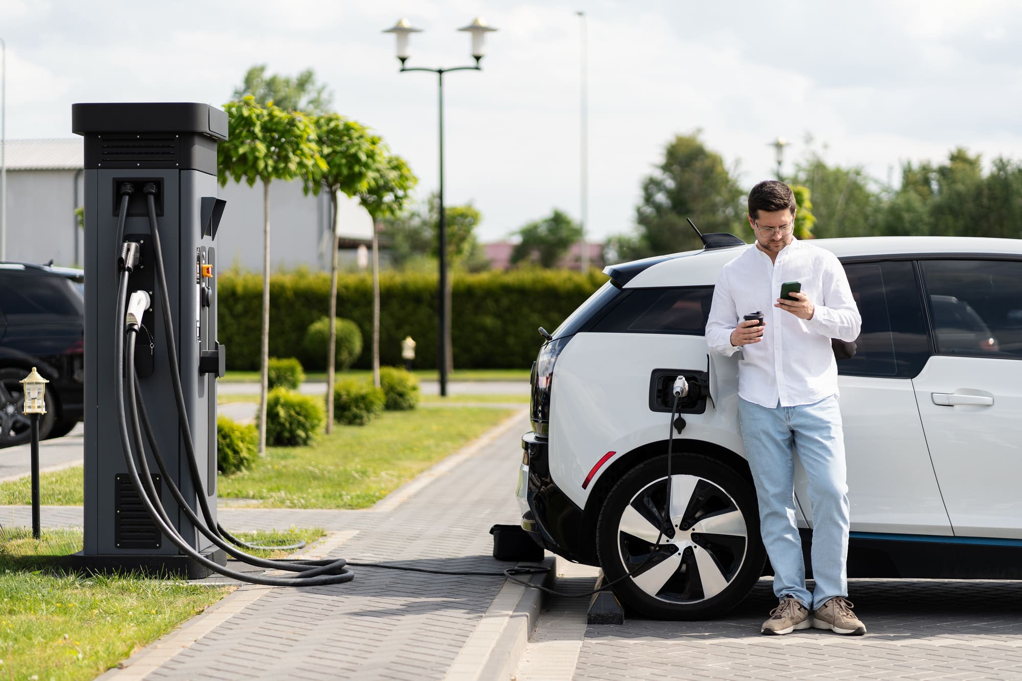 Man charging electric car at charging station and using smartphone in urban area