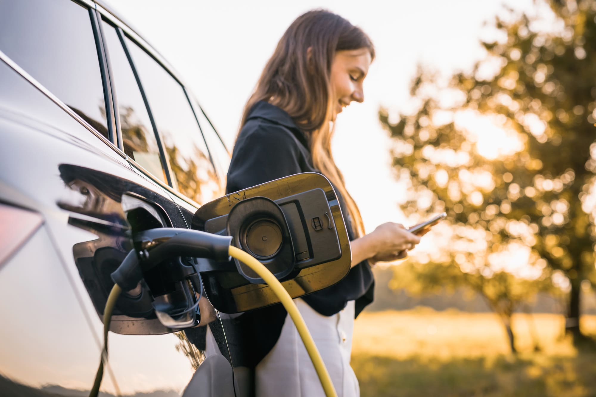 A young woman plugs a charger into her electric car, her smartphone in hand using the ChargeHub application.