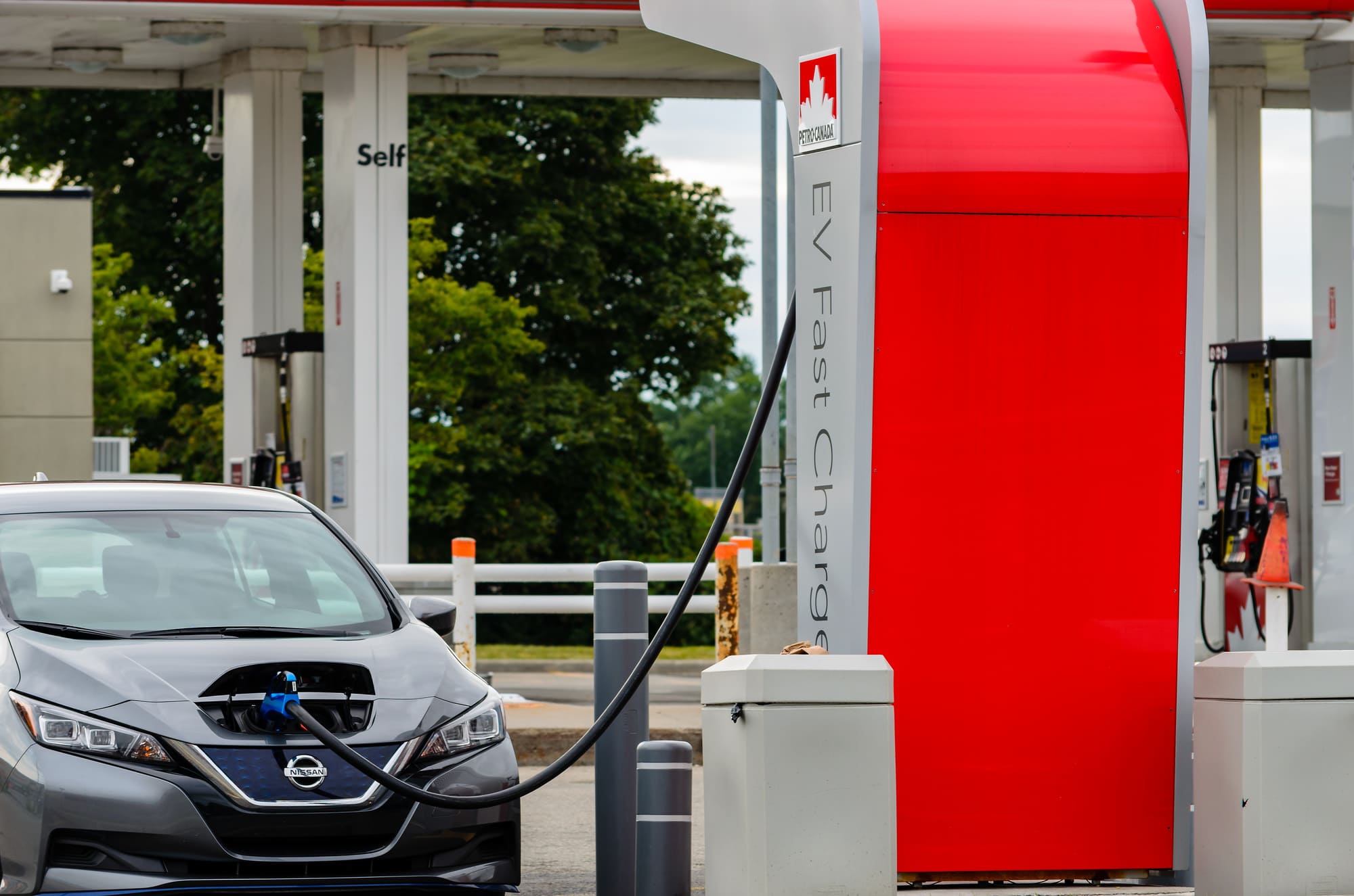 Electric car plugged into a fast-charging station, cable connected to the front of the vehicle at a charging station.